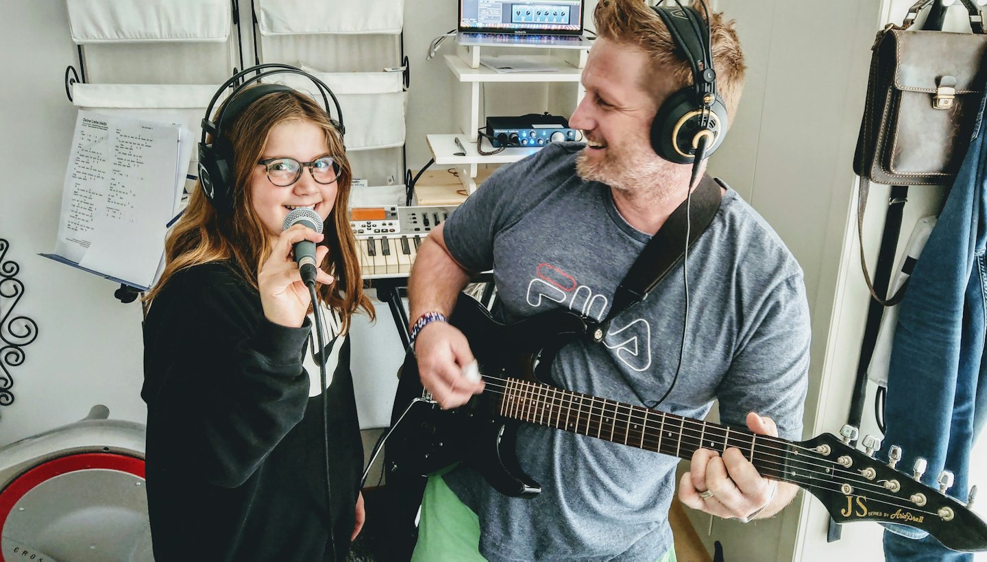 Uomo suona la chitarra elettrica mentre una ragazza canta al microfono in uno studio casalingo.